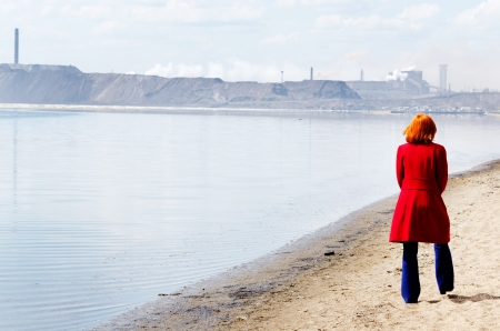 Young woman walks alone on a beach heading towards industrial landscapeの写真素材