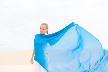 Young slim woman wearing white with flying blue scarf in desertの写真素材