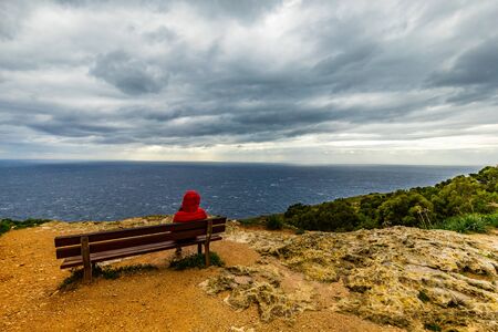 bench on the sea cliff on Malta with red jacket hooded woman on a cloudy storm day