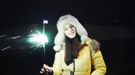Young cute girl in a winter fur hat and yellow jacket in a festive mood. Sparkling burning sparklers in hand with flying sparks. Dancing and having fun. Holidays conceptの写真素材
