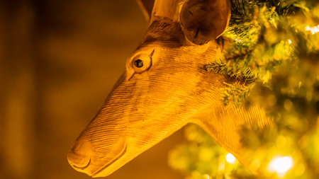Deer head with Christmas garlands and fairy lights on wall closeup. Toy animal decorated with fir twigs and lights on blurry background. Cute Xmas holiday ornament in semi-dark roomの写真素材
