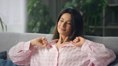 Woman in her 30s or 40s enjoys a peaceful moment at home, sitting on a sofa, stretching her arms,...