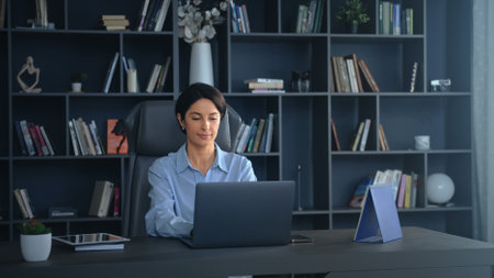 Confident businesswoman working on laptop in modern office with bookshelf. Focused and successful, typing away, embodying a dedicated career womanの写真素材