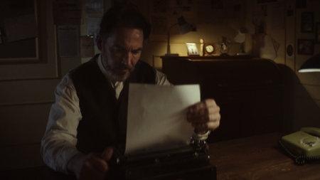Focused male writer working on a vintage typewriter in his dimly lit office at night, holding a...