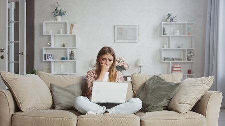 Young woman sitting on the sofa with crossed legs is covering her mouth with her hands and crying, reading bad news on a laptop, having a mental breakdownの写真素材