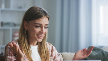 Happy young woman using laptop sitting on comfortable sofa, gesturing and smiling while video chatting with friends or family in living roomの写真素材