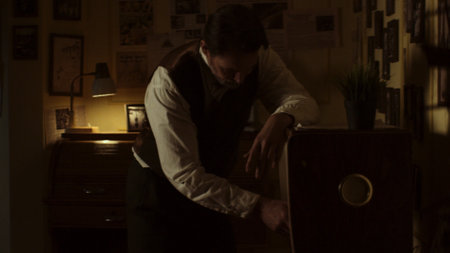 Writer is working late at night in his office, illuminated by a desk lamp, searching through files in a vintage wooden cabinet, surrounded by pictures and documents on the wallの写真素材
