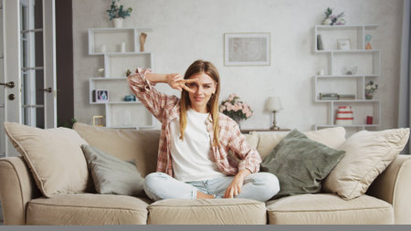 Cheerful young woman sitting with crossed legs on comfortable sofa in cozy living room, making victory gesture with fingers near eye and smiling, enjoying weekend at homeの写真素材