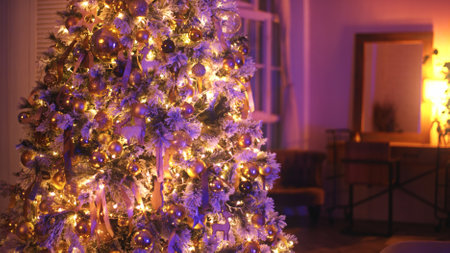 Christmas tree decorated with golden baubles, ribbons and fairy lights is illuminating a cozy living room with purple lightsの写真素材
