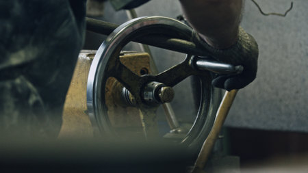 Close up of a metalworker's gloved hand operating a vintage industrial machine, turning a lever attached to a rotating wheel, possibly involved in metal shaping or processingの写真素材
