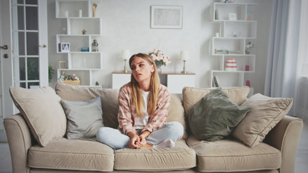 Sad woman is sitting on the sofa in the lotus position in her living room, feeling depressed, lonely and disappointed, she is suffering from psychological problemsの写真素材