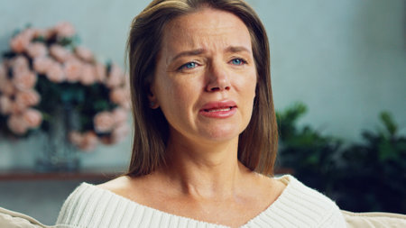 Mature woman crying with tears streaming down her face, expressing deep sadness and emotional distress while sitting on a sofa in a living room with blurred flowersの写真素材