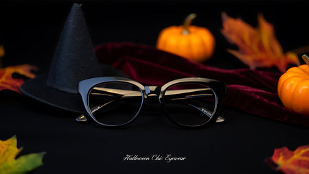 A still life composition with a pair of black, stylish eyeglasses placed in the foreground. Behind the glasses, a black witch hat, a piece of dark red velvet fabric, and several orange pumpkins and autumn leaves are arranged against a dark background. The overall theme is Halloween chic.の素材