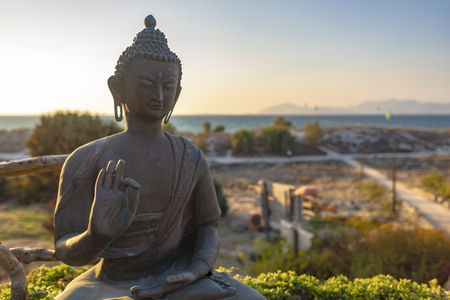 Sunset over the representation of the sitting protection Buddha hand gesture and posture - with right hand raised, facing outward, left hand resting in lap, and seated with one leg resting on top of the other (Single Lotus) - with sea and beach as a background.の写真素材
