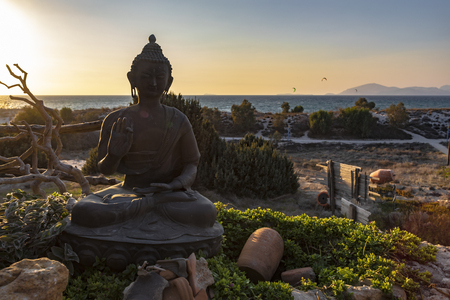 Sunset over the representation of the sitting protection Buddha hand gesture and posture - with right hand raised, facing outward, left hand resting in lap, and seated with one leg resting on top of the other (Single Lotus) - with sea and beach as a background.の写真素材