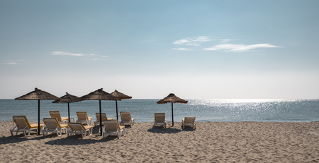 Landscape view of lounge relaxation chairs and straw made umbrellas on a sandy beach in front of the horizon line over turquoise blue sea.の写真素材