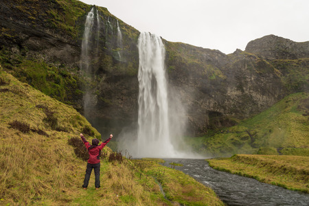 Happy woman by waterfall Skogafoss on Iceland posing serene and free outdoors with arms raised. Girl visiting famous tourist attractions and landmarks in Icelandic nature landscape, ring road.の写真素材