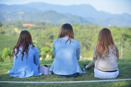 Three young women viewed from behind while wearing casual dress and sit on green grass in garden.の写真素材