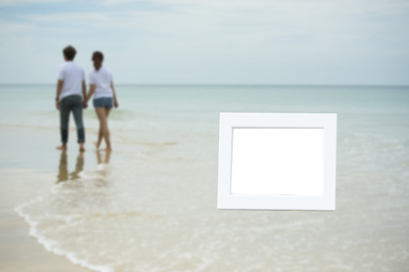 blurred of young asian couple hold hand walk together on sea sand beach in left coner with white empty picture frame.の写真素材