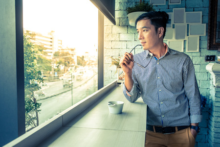 Young Asian man hand hold eye glasses looking out of window, Casual professional entrepreneur working-in out of office in cafe or coffee shop, modern lifestyle and digital age conceptsの写真素材