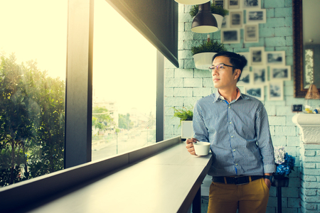 Young Asian happy man look through clear window and thinking during coffee time in cafe, casual freelance lifestyle or digital nomad conceptsの写真素材