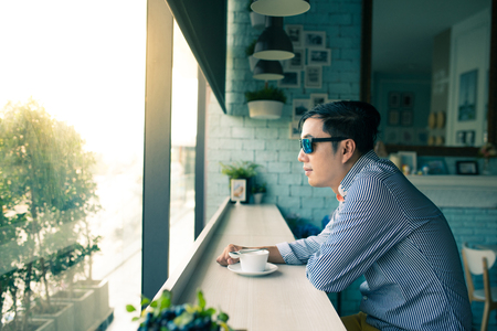 Young Asian happy man look through clear window and thinking during coffee time in cafe, casual freelance lifestyle or digital nomad conceptsの写真素材