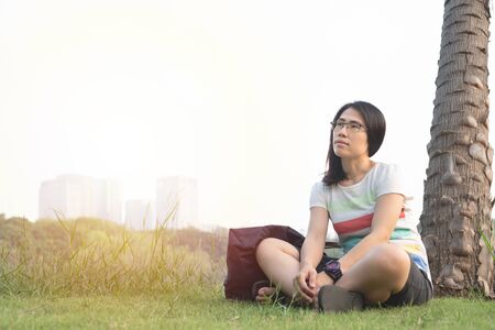 Portrait of asian woman wear glasses and casual dress sit on green lawn in public park look up the sky with happy face.の写真素材