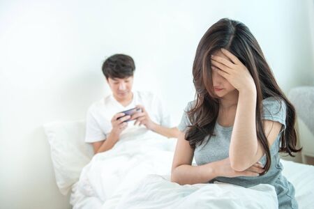 Young asian woman is upset that man uses phone all the time without paying attention to her. Man holds phone pay attention on smart mobile phone while sit on bed with ignoring on his girlfriend.の写真素材