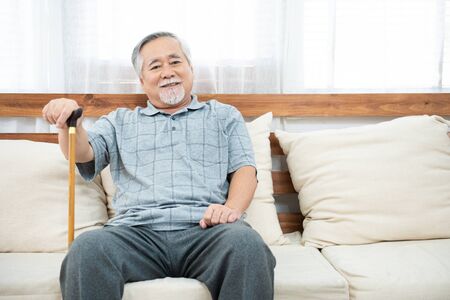 elderly old senior man sitting, resting his hands on wooden walking stick sitting on couch in living...