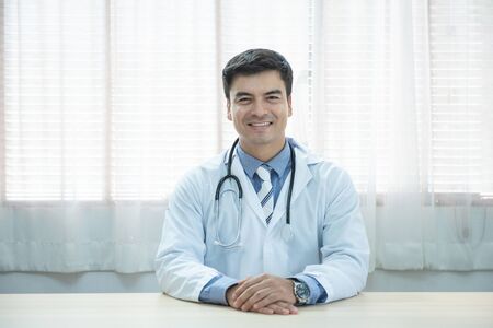 Young doctor caucasian man sitting at the desk at his working place and smiling looking at camera. Perfect medical service in clinic. Happy future of medicine and healthcare.の写真素材