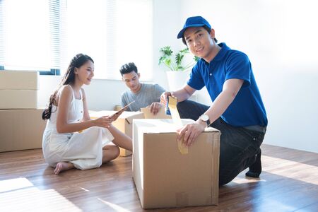 Delivery service worker man in blue uniform help happy couple customer packing parcel with tape in...