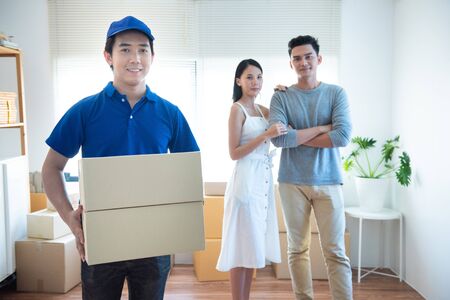 Smiling young asian delivery man in blue uniform holding and carrying two cardbox parcel look at camera with happy young asia couple sender standing in background.の写真素材