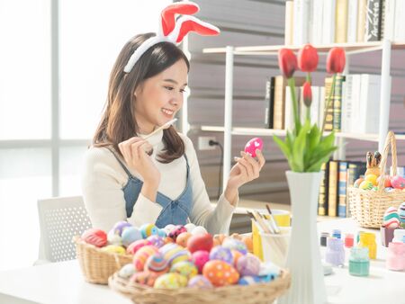 Beautiful young asian woman painting easter eggs on table with decorated eggs for celebrate April...