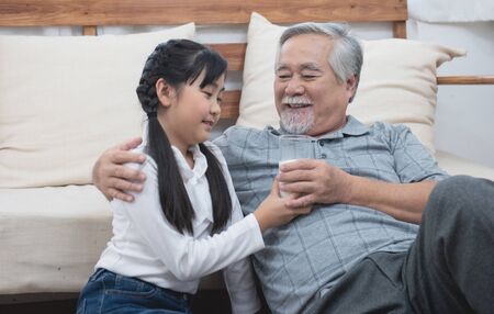 Happy asian senior elderly grandfather have grandchild look after and take care with giving milk and kiss on cheek while sitting on sofa at home,retirement health lifestyle concept.の写真素材