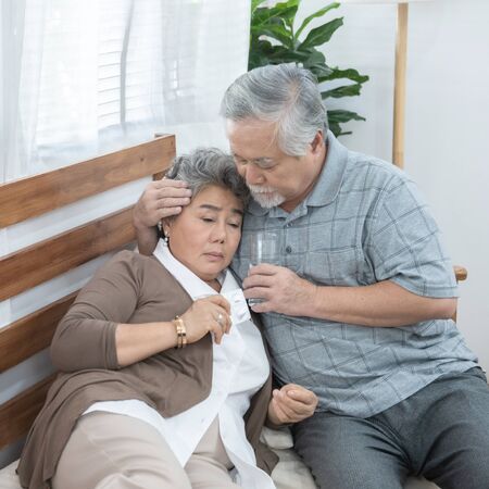 Asian Senior Woman taking medicines and drinking water while sitting on couch. Old man take care his wife while her illness at the house.の写真素材
