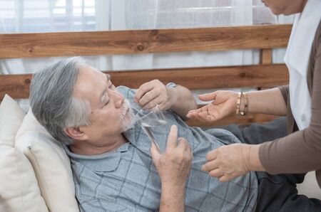 Asian Senior man taking medicines and drinking water while lie on couch. Old woman take care his...