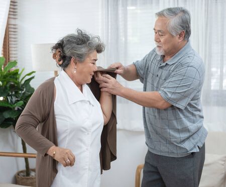 Asian senior elder man help elder woman wear a shirt at home.の写真素材