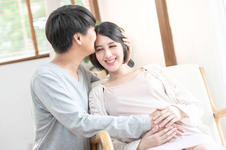 Happy excited pregnant couple having fun at home. Cheerful expectant mom sitting on couch dad kiss forehead, look out window smiling, laughing. Enjoying pregnancy motherâs day conceptの写真素材