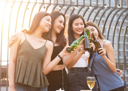 Group of young beautiful happy asian women holding bottle of beer chat together with friends while celebrating dance party on outdoor rooftop nightclub with copy space for advertising.の写真素材