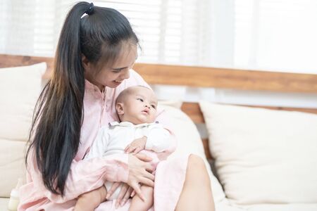 family, child and parenthood concept - happy beautiful young asian mother smiling hugging holding newborn baby in her arms at home. Motherâs day banner with copy space for advertiseの写真素材
