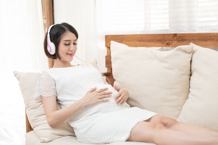 Relaxation in pregnancy. Calm pregnant young asian girl Listen to music Mozart effect good for fetus by using headphones attached to the stomach hold earphones near belly lying on sofa at home.の写真素材