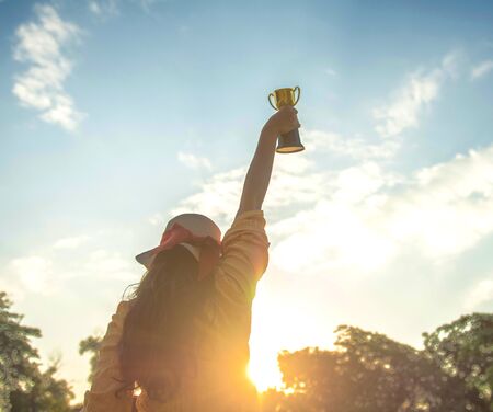 Back or rear view of long hair women hands hold gold trophy cup on blue sky with cloud and sunrise background, for symbol of victory and success for business concept.の写真素材