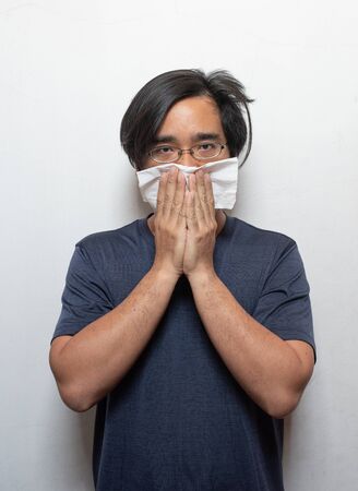Asian man getting cold or flu,he is coughing to clear throat with hand holding tissue paper closing his mouth isolated on white background Illness and sickness concept.の写真素材