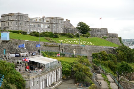PLYMOUTH, UK - JULY 4 2016: Tourists sit in an outdoor cafe and walk along the public promenade in the British south coast seaport city of Plymouth, Devon. The Royal Citadel and a grass sign reading Welcome to Plymouth are in the background.のeditorial素材