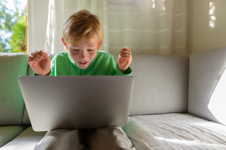 Happy boy smiling while watching on laptop and sitting on sofa at homeの写真素材