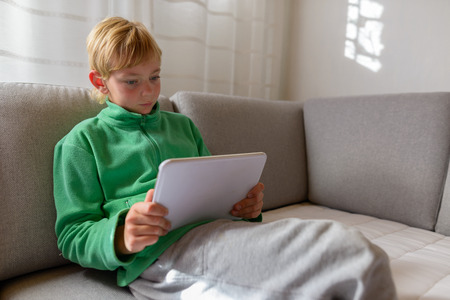 Handsome boy using digital tablet and sitting on sofa at homeの写真素材