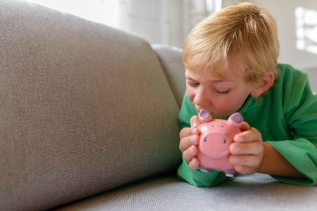 Handsome boy holding and peeking at piggy bank while lying on sofa at homeの写真素材