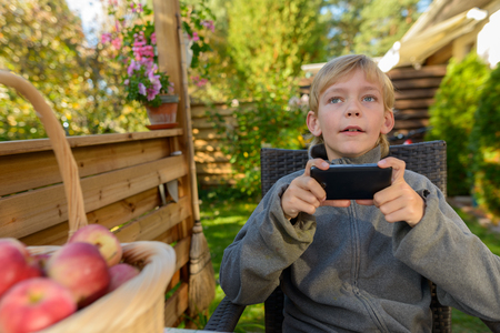 Handsome boy holding mobile phone while thinking in the backyard with basket full of apples at homeの写真素材
