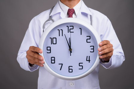 Hands of young Indian man doctor holding wall clock against grayの写真素材