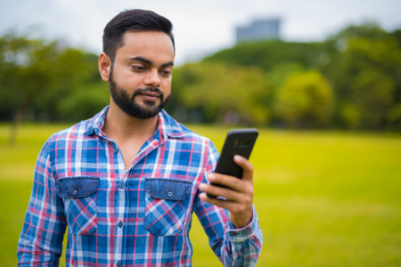 Portrait Of Young Handsome Indian Man In Park Using Mobile Phoneの写真素材